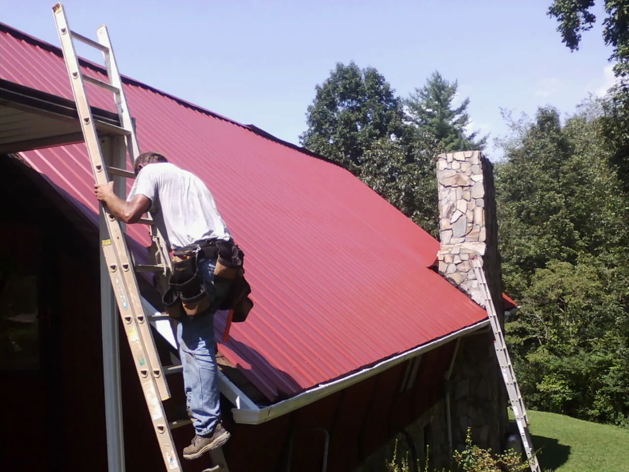 Roofer on a ladder beside a red metal roof and stone chimney — Soffit & Fascia Repair by Roofing Brothers in Sloatsburg